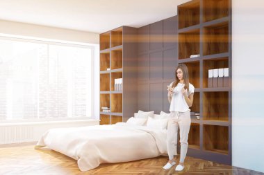 Bir ahşap zemin üzerinde kitaplıklar arasında beyaz yatak duran bir beyaz ve gri duvar yatak köşede bir akıllı telefon olan kadın. 3D render sahte kadar çift tonlu görüntü pozlaması