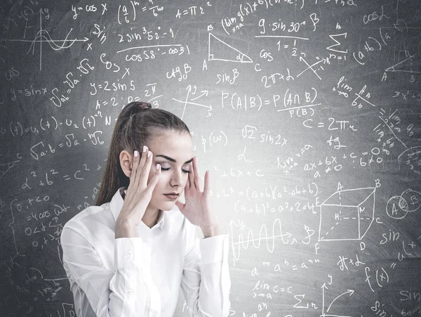 Stressed young businesswoman with long hair in a ponytail wearing a white shirt. She is having a headache and standing with closed eyes. A blackboard background with a physics formulae on it.