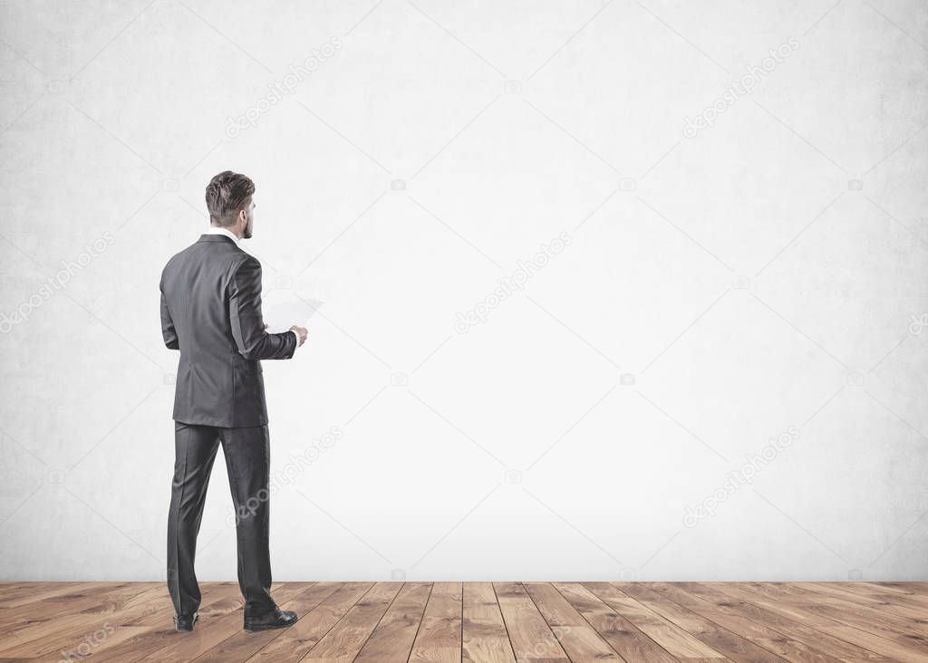 Young businessman with a beard wearing a gray suit is standing and holding papers. He is looking forward. Concept of leadership. A mock up concrete wall background