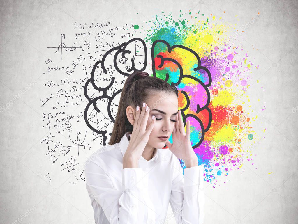 Stressed young businesswoman with long hair in a ponytail wearing a white shirt. She is having a headache and standing with closed eyes. A concrete wall with a colorful brain sketch