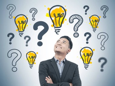 Confident and cheerful Asian start up founder in suit standing near a gray wall with many question marks and light bubls. clipart