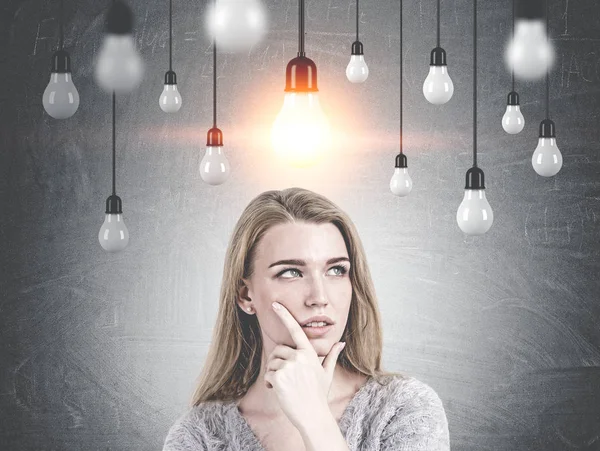 Head and shoulders portrait of a gorgeous young woman with gray eyes and blond hair looking into distance and thinking. Yellow light bulbs near a blackboard. An idea concept