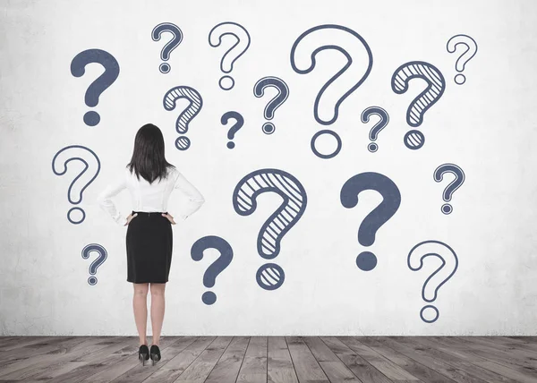 Elleri beli üzerinde duran ve ileriye dönük uzun siyah saçlı Genç bir iş kadını arkadan görünüş. Birçok soru işaretleri bir beton duvar.