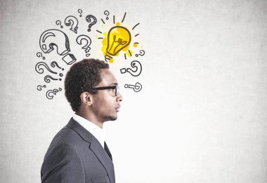 Gözlük ve gri takım elbise giyen genç yakışıklı Afrikalı Amerikalı bir iş adamını yan görünüm. Birçok soru işaretleri bir beton duvar ile parlak renkli ışık ampul. Yukarı alay