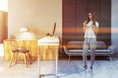 Bir modern İskandinav tarzı ev ofis bir dar bilgisayar masa ve şeffaf bir saç bir beton zemin üzerinde rahat gri kanepede duran bir telefon olan kadın. 3D render tonlu görüntü