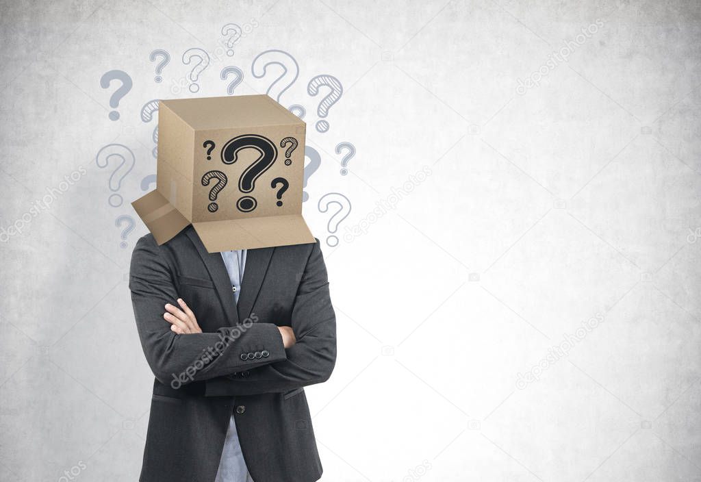 Portrait of a young businessman wearing a dark suit and standing with crossed arms with a box on his head. A concrete wall background. Question marks around the head. Mock up