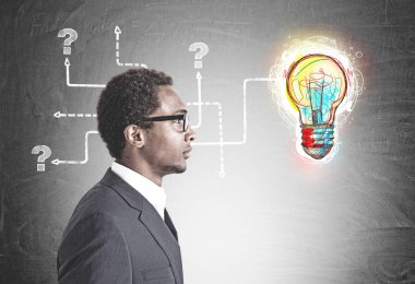 Side view of a young handsome African American businessman wearing glasses and a gray suit. A bright colorful light bulb with tangled arrows drawn on a blackboard clipart