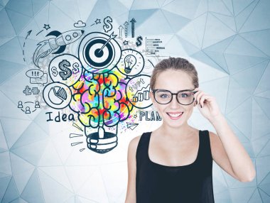 Neşeli genç kadın gözlüklerini dokunmaktan ve kameraya bakıyor. Siyah kolsuz bluz ve uzun saçları at kuyruğu ya da topuz. Bir ampul ve iş ikonları/simgeleri ile desen gri bir duvar içinde renkli beyin.