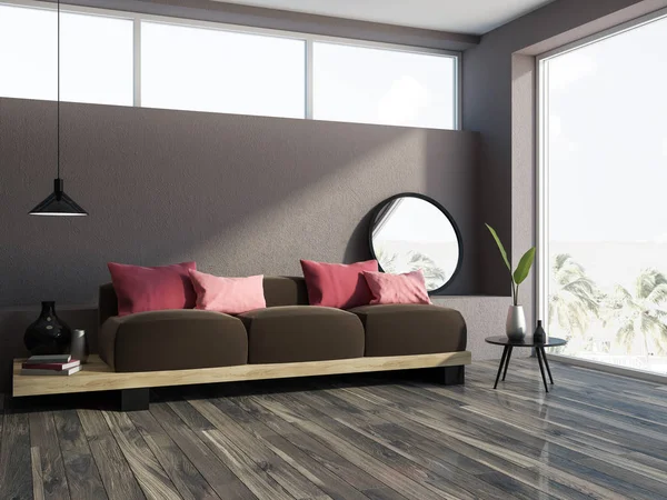 Brown living room corner with a wooden floor, a loft window and a long comfortable dark brown sofa with red cushions on it. 3d rendering mock up