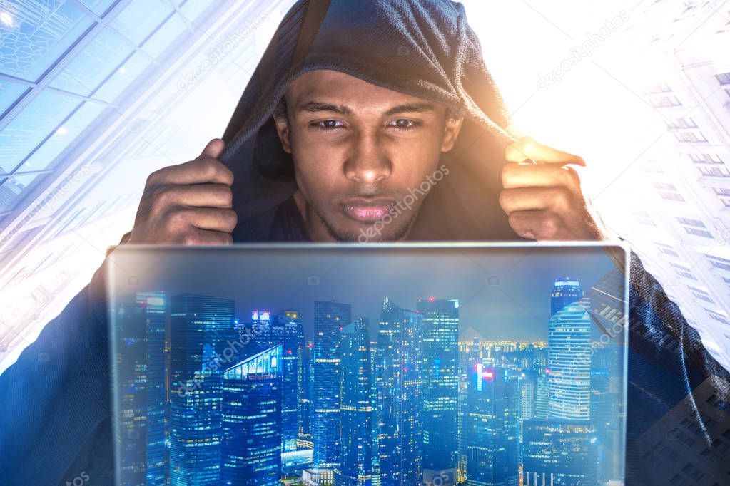 Young African American hacker in a black hoodie looking at his laptop screen. Double exposure of a night city. Morning cityscape background. Toned image