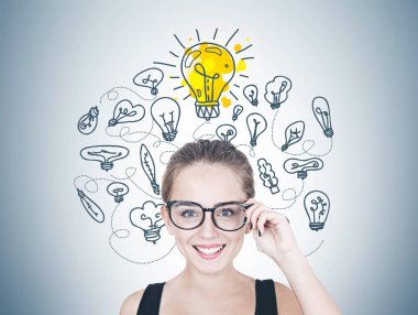 Gözlüklerini dokunmaktan ve kameraya bakarak neşeli bir genç kadın portresi. Siyah kolsuz bluz ve uzun saçları at kuyruğu ya da topuz. Birçok ışık ampuller ile gri duvar. Bir parlak parlıyor