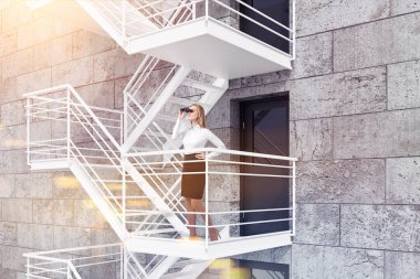 Modern bina dış beton tuğla duvarlar, Gri kapı ve acil çıkış merdivenleri ile sarışın işkadını dürbün ile yan görünüm. 3D render sahte kadar çift tonlu görüntü pozlaması