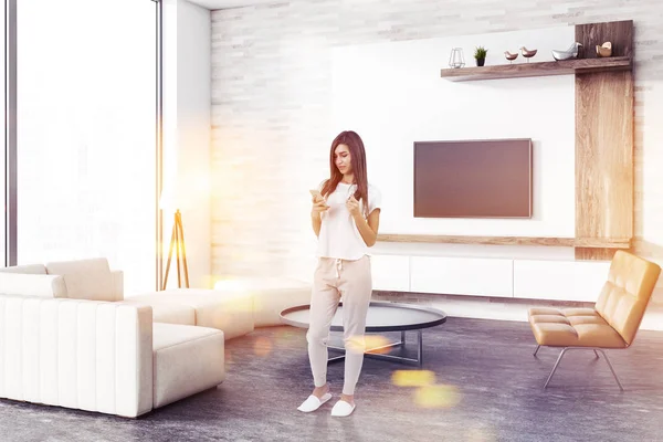 Woman with smartphone in loft living room with concrete floor, white and wooden walls, a sofa and a leather bench. Round coffee table and a flat TV set on the wall. 3d rendering mock up toned image
