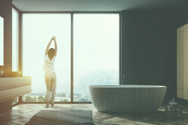 Gri şık banyo ahşap zemin, gri halı, küvet ve lavabo dikey ayna ile büyük penceresinde arayan kadın. Kopya alanı görüntü çift pozlama gökdelen tonda