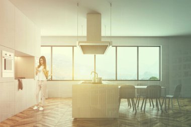 Beyaz duvarları, ahşap zemin, büyük pencereler, beyaz ada ve mutfak tezgahı ile modern mutfak ve yemek odası masa sandalye ile bir genç kadın. Çift tonlu görüntü pozlaması kadar alay