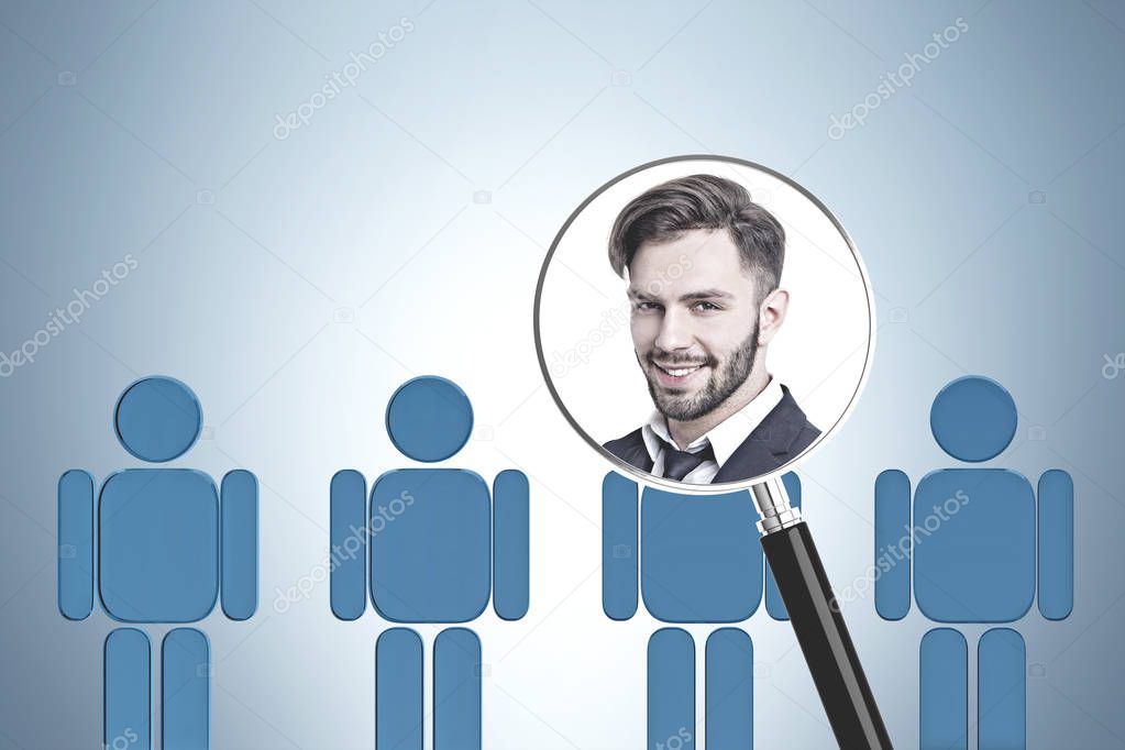 Portrait of smiling young businessman in magnifying glass over row of blue human figures in gray room. HR concept.