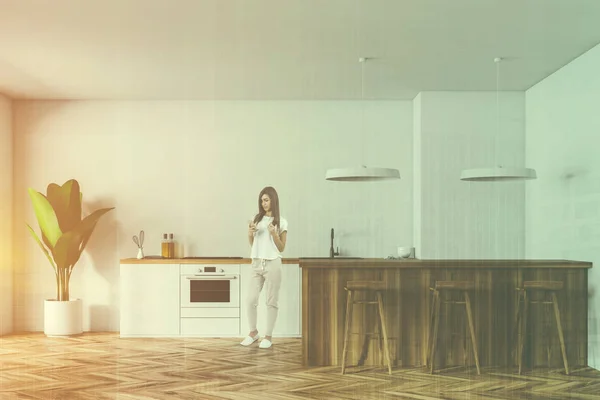 Young woman with smartphone in modern kitchen with white walls, wooden floor, white countertops, a wooden bar with stools and big plant in a pot. Copy space toned image double exposure