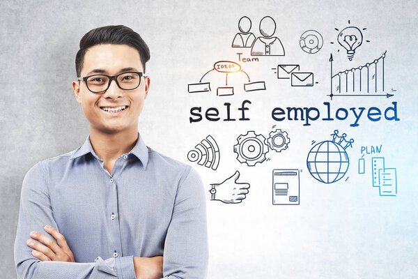 Smiling Asian man in glasses and shirt standing with crossed arms near a concrete wall with self employed text and icons on it. Toned image