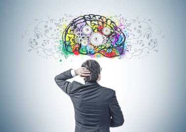 Bir koyu takım elbise ve onun baş iş planı kroki ve beyin gears ile gri duvara bakarak tırmalamak siyah saçlı Genç beyaz işadamı arkadan görünüş