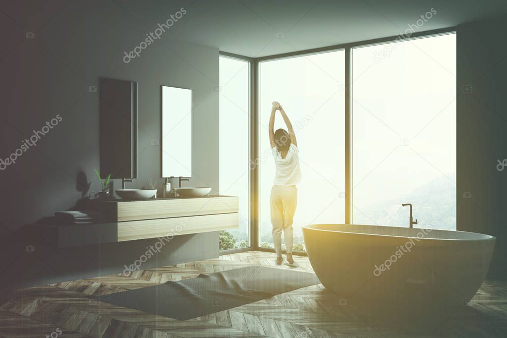 Woman in pajamas in gray bathroom interior with wooden floor, gray rug, window with forest, bathtub, and double sink with mirrors. 3d rendering copy space toned image double exposure skyscraper