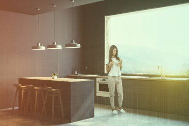 Smartphone ile beton zemin gri mutfak köşesi, dağ manzarası ve gri mutfak tezgahı ile büyük pencere içinde olan genç kadın. Bar tabureleri ile. Çift tonlu görüntü pozlaması