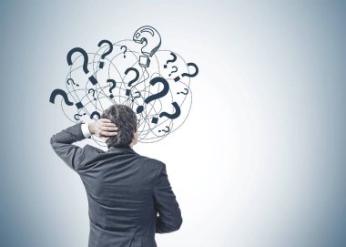 Bir koyu takım elbise ve soru işaretleri gri duvara bakarak başını tırmalamak siyah saçlı Genç beyaz işadamı görünümünü arka. Kopya alanı