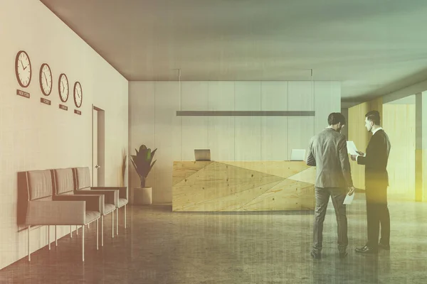 Two business partners discussing company issues in office hall with wooden reception desk, clocks and glass walls. Toned image double exposure