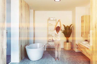 Lüks banyo beton zemin, çift lavabo ahşap ayakta ve tezgah, iç raflar Rulo havlu ve kremler ve beyaz küvet ile. Genç Kadın pijama. Tonlu görüntü