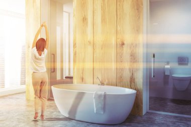 Yan görünüm ahşap duvarlar, beyaz lüks banyo fayans zemin ve beyaz küvet. Dikey ayna ve bir tuvalet. Pijamalar içinde duran genç kadın. Tonlu görüntü