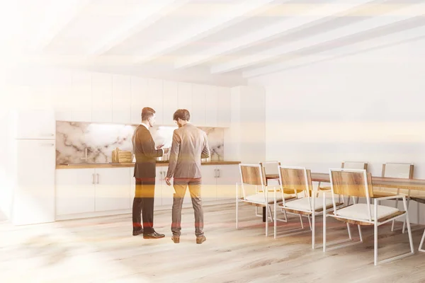 Two men talking in white kitchen interior with wooden floor, white bridge and countertops with built in appliances and white marble wall fragment. Long table with white chairs. Toned image