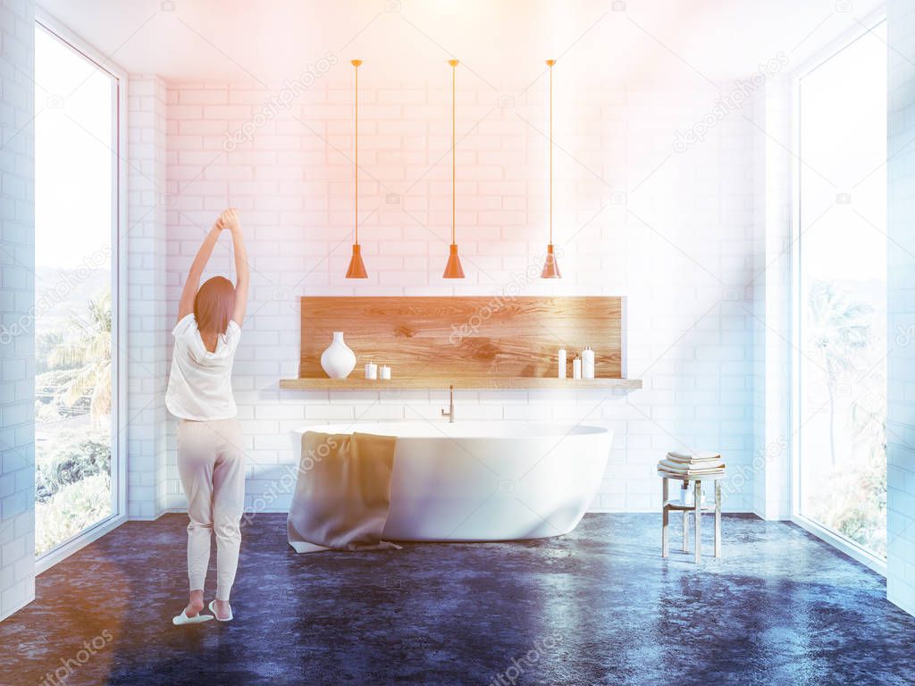 Woman stretching in the morning in white brick bathroom interior with a concrete floor, a white bathtub, several ceiling lamps and a wooden shelf with candles. Toned image