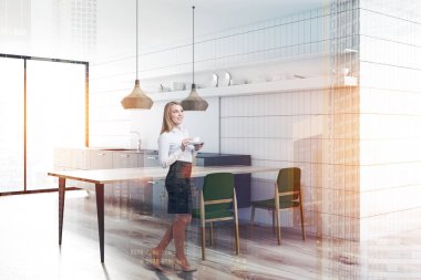 Sarışın kadın kahve iç beyaz çini duvarlar, gri mutfak tezgahı, yeşil sandalye ve şık tavan lambaları ile uzun ahşap masa ile modern mutfak ile. Çift tonlu görüntü pozlaması