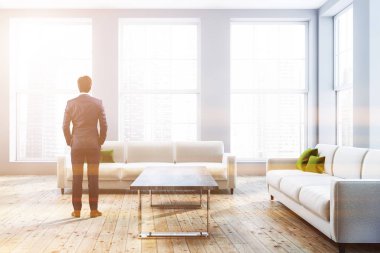 Gri duvarlar, ahşap zemin, yeşil yastıkları ile iki beyaz koltuklar ve sehpa ile lüks oturma odasında duran genç işadamı görünümünü arka. Tonlu görüntü