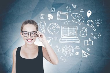 Siyah kolsuz bluz ve Internet simgeleri ile geometrik desenli koyu gri duvarının yakınında duran gözlük gülümseyen genç kadın. Merhaba teknoloji kavramı.