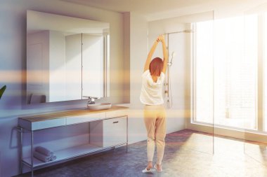 Modern banyo köşesinde beyaz duvarlar, beton zemin, lavabo beyaz tezgah ve büyük ayna üzerinde ayakta duran kadın. Cam kapı duş. Tonlu görüntü