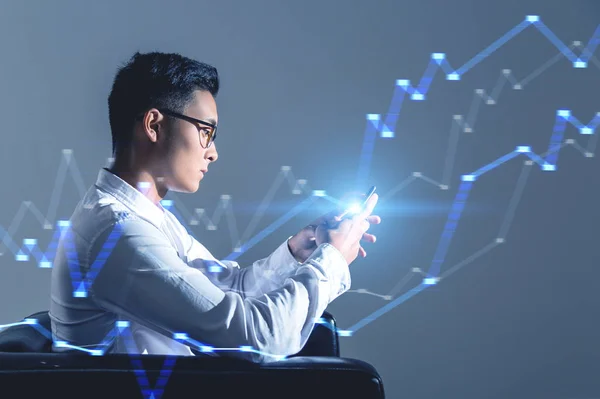 Asian businessman sitting in armchair with smartphone near gray wall. Glowing graphs in the air. Toned image double exposure