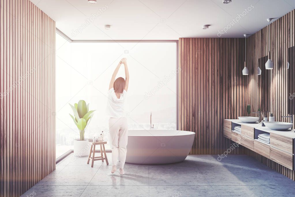 Woman in pajamas in bathroom interior with wooden walls, gray floor, white bathtub and double sink. Panoramic window. Toned image