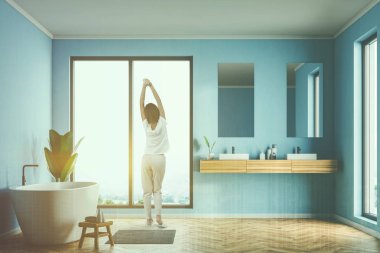 Loft banyo iç mavi duvarlar, ahşap zemin, beyaz küvet ve iki dikey aynalar ile çift lavabo ile duran kadın. Çift tonlu görüntü pozlaması
