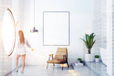 Modern banyo beyaz duvarlar, beton zemin, beyaz küvet ve kahverengi koltuk üstünde asılı poster kadar dikey sahte ile duran kadın. Yuvarlak pencere. Çift tonlu görüntü pozlaması