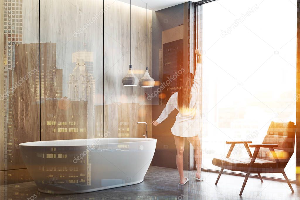 Woman standing in bathroom corner with wooden walls, concrete floor, white bathtub and leather armchair standing near loft window. Toned image double exposure