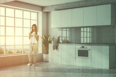 Minimalist mutfak iç beyaz duvarlar, beton zemin, beyaz mutfak tezgahı ve dolapları ve büyük pencere ile smartphone ayakta olan kadın. Çift tonlu görüntü pozlaması