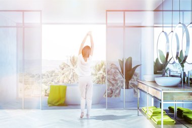 Kadın lüks banyo iç ile beyaz duvarlar ve zemin, çift lavabo ahşap tezgah, beyaz küvet ve tropik manzaralı çatı penceresi tutuyoruz. Tonlu görüntü