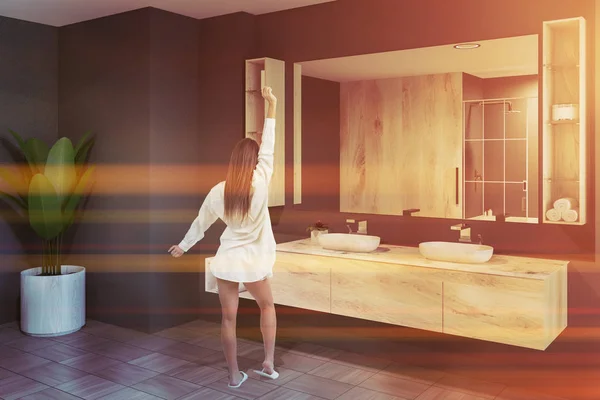 Woman in nightgown in modern bathroom interior with gray walls, tiled floor and white double sink standing on wooden countertop with big mirror hanging above it. Toned image