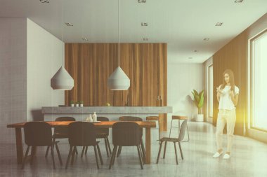 Kadın ile smartphone beyaz ve ahşap duvarlar ile modern dining room, koyu gri sandalye ve büyük pencereler ile ahşap masa. Tonlu görüntü