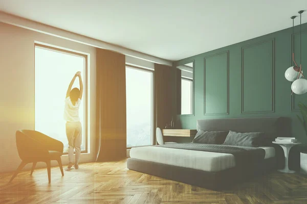 Woman standing in modern bedroom with green walls, wooden floor, gray master bed and make up table in the corner. Black armchair near the window. Toned image double exposure