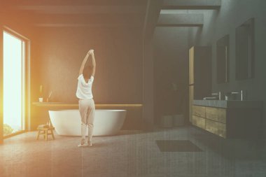Gri duvarlar ve zemin, çatı penceresi, beyaz küvet ve ahşap tezgah üzerinde duran mermer çift lavabo banyo içinde duran kadın. Çift tonlu görüntü pozlaması