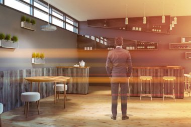 Modern bar iç Gri duvarlar, ahşap masa, küçük beyaz sandalye ve gri bar tabureleri ile ahşap yuvarlak ile ayakta adam. Şişe raflarda. Tonlu görüntü