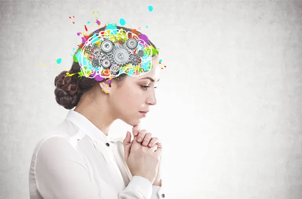 Side view of pensive young businesswoman with dark hair wearing white blouse with colorful brain with gears inside her head. Concept of decision making. Concrete wall background. Mock up