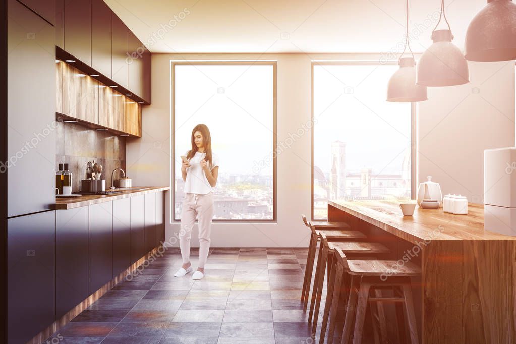 Woman standing in loft kitchen with gray walls, gray and wooden countertops and bar with stools and coffee machine. Toned image