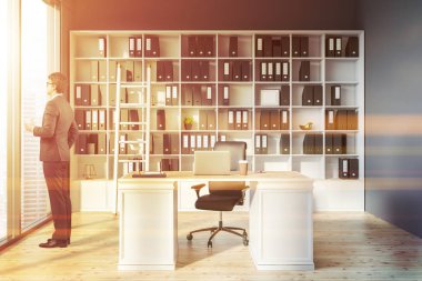 Adam Yöneticisi office iç ray duvarlar, ahşap zemin, panoramik pencere, dizüstü bilgisayar ve kitaplık klasörlerle beyaz masa ile ayakta. Tonlu görüntü